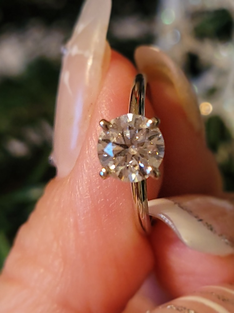 Customer photo of a four-prong solitaire engagement ring with a round diamond, held between two fingers